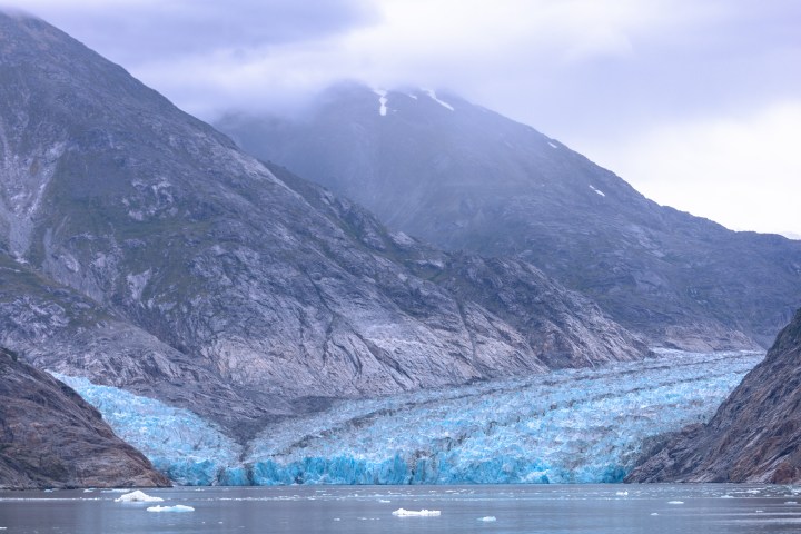 Glacier flowing between mountains with cloudy sky and water below.