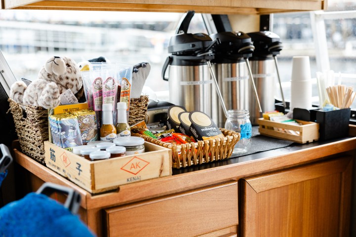 Snack and beverage station with plush toys, snacks, sauces, and hot drink dispensers on a wooden counter.
