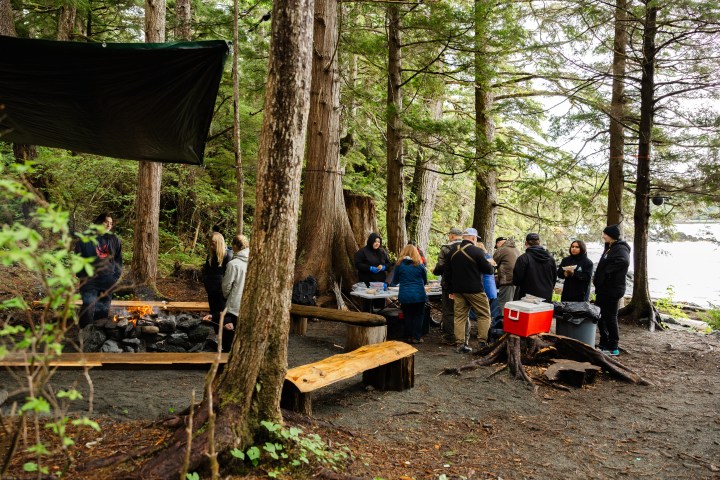 Group of people gathered in a forested area with a campfire under a tarp shelter.
