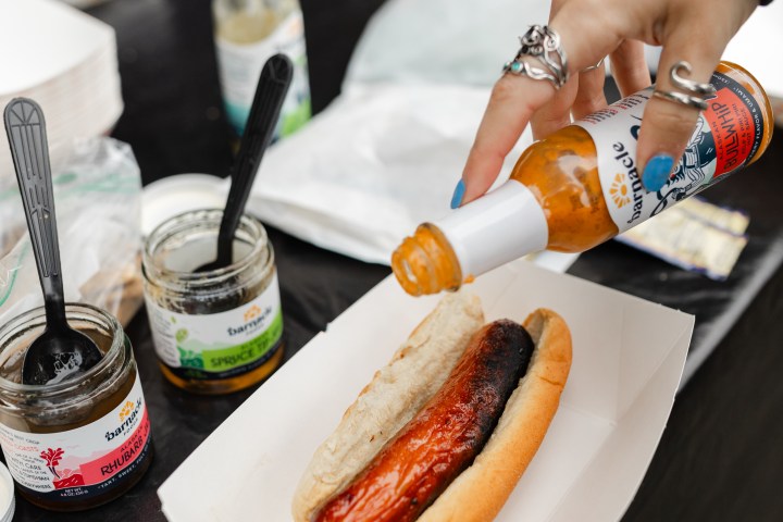 Hand pouring sauce on sausage in a bun, next to condiments on a table.