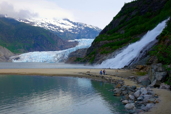 A glacier with snowy mountains, a waterfall, and people walking near a lake.