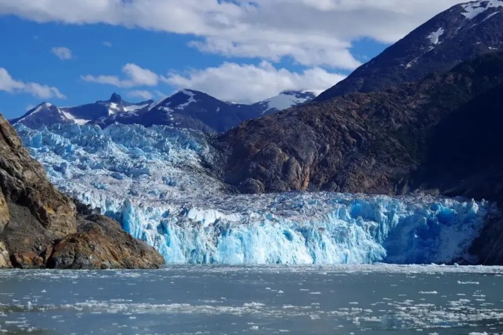 Blue glacier surrounded by mountains and a lake under a partly cloudy sky.