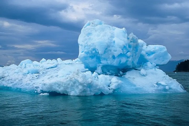 Large blue iceberg floating in calm ocean under cloudy sky.