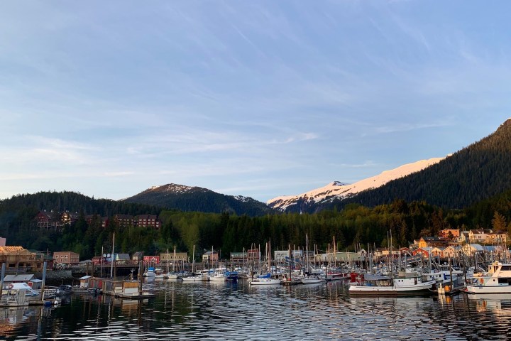 Scenic harbor with boats, mountains, and buildings under a clear sky at dusk.