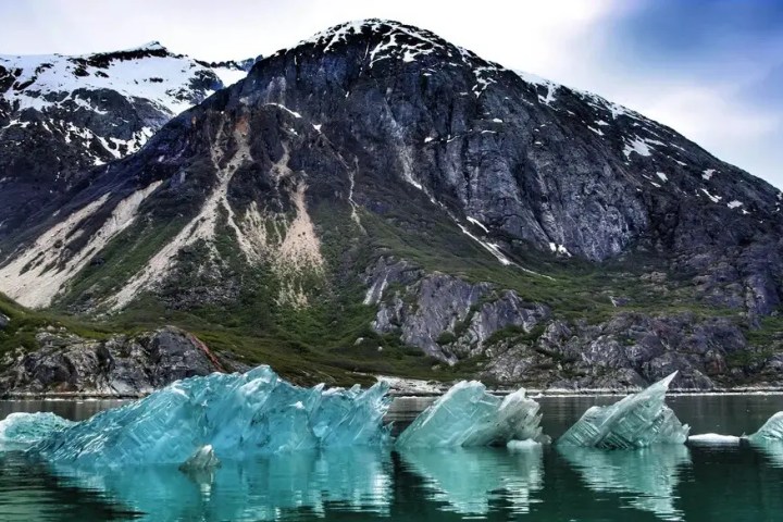 Jagged blue icebergs floating in front of a rocky, snow-peaked mountain.